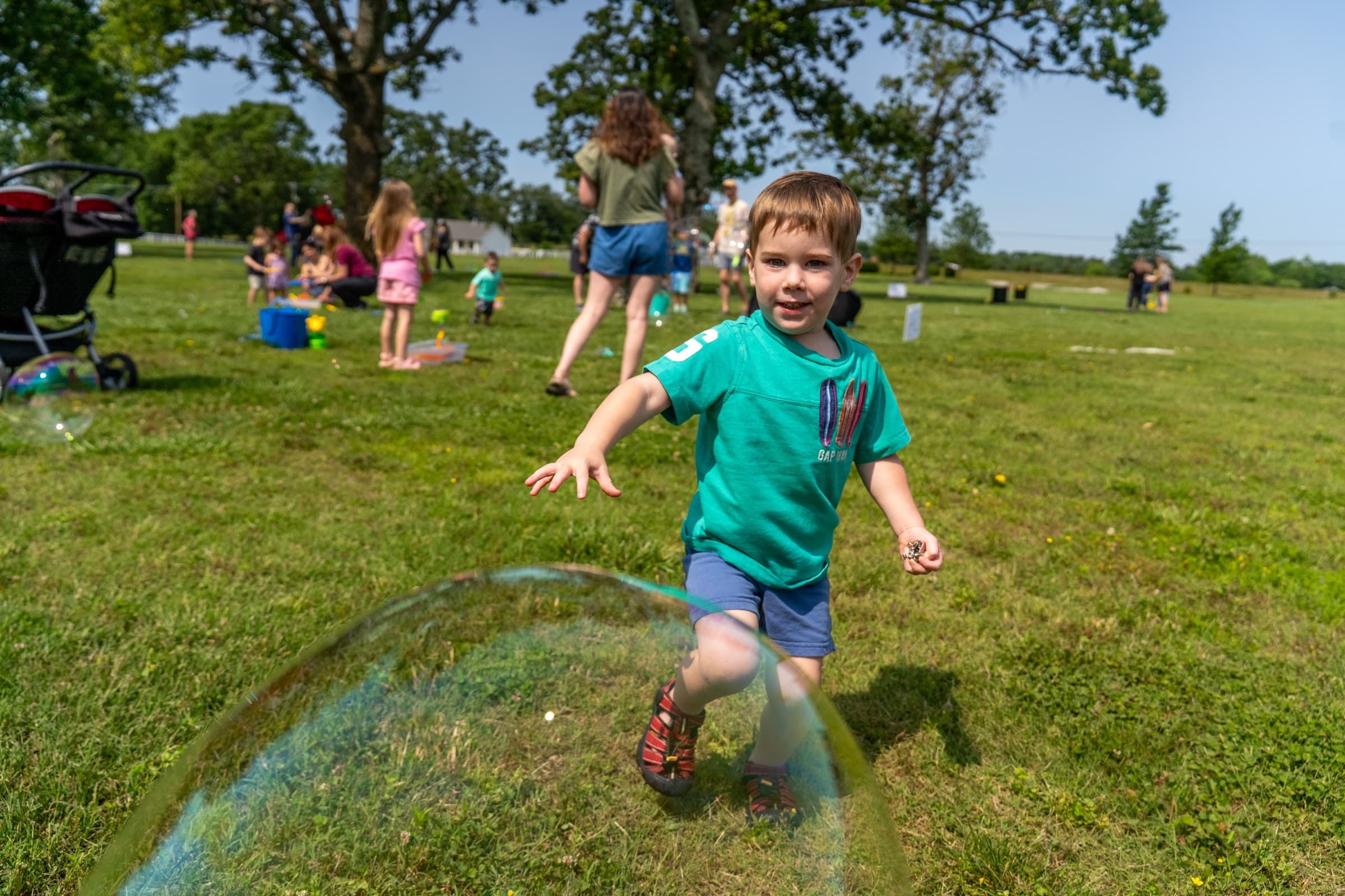 A child runs through a giant soap bubble on the grass at an Inclusive Connections community event, with families gathered in the background.