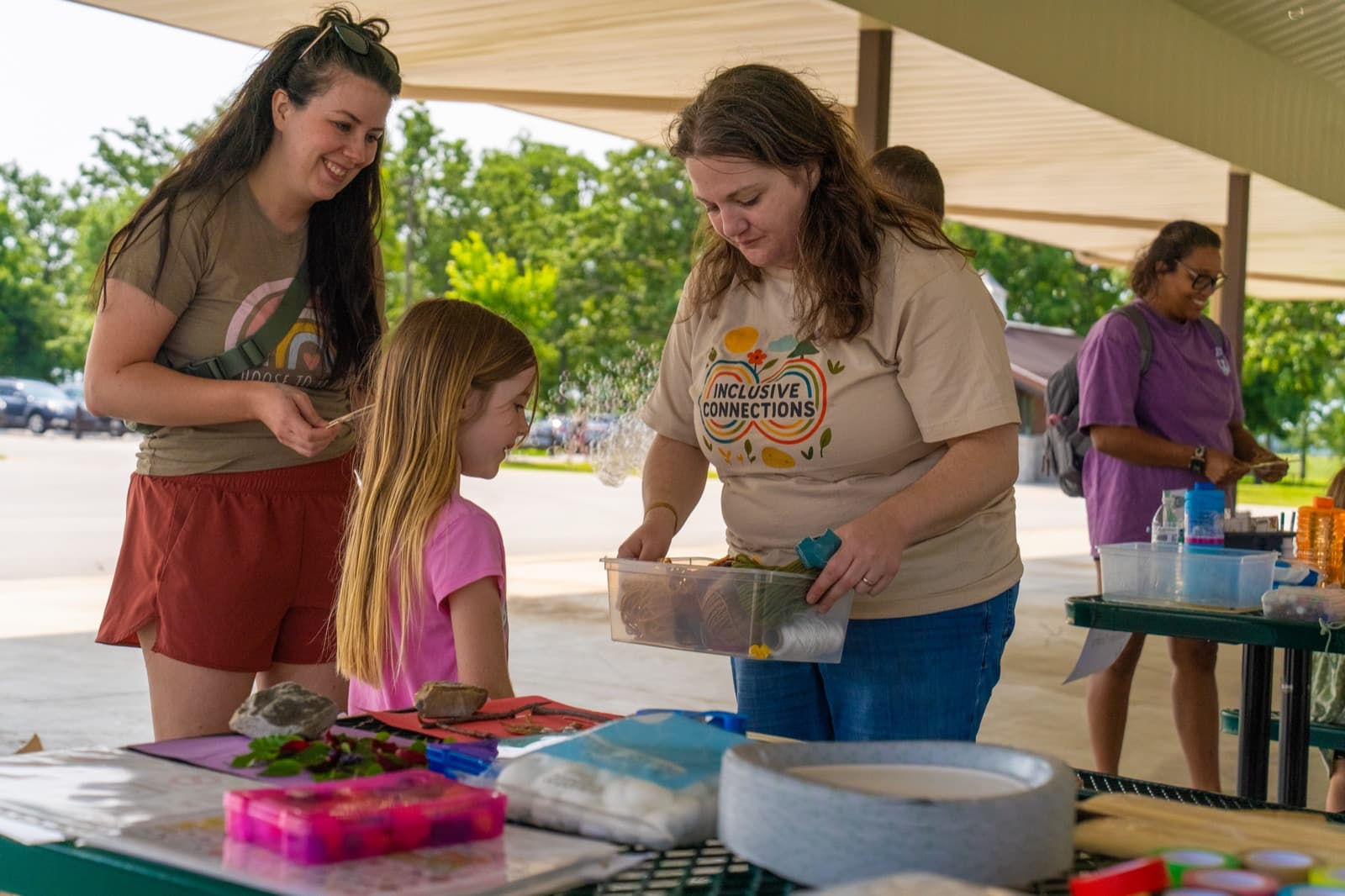 Volunteers set up a craft activity table under a pavilion while a child looks on.
